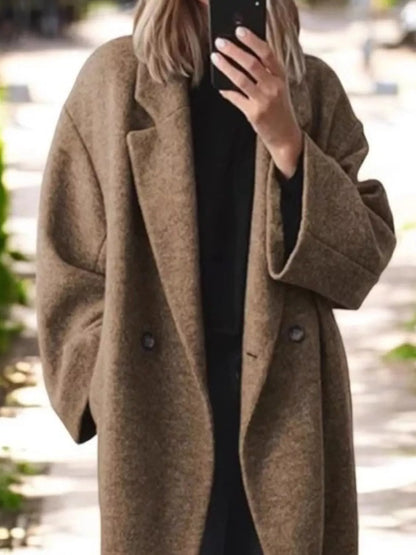 A person outdoors wearing an oversized brown wool-blend coat over a black outfit, holding a phone to take a mirror selfie.