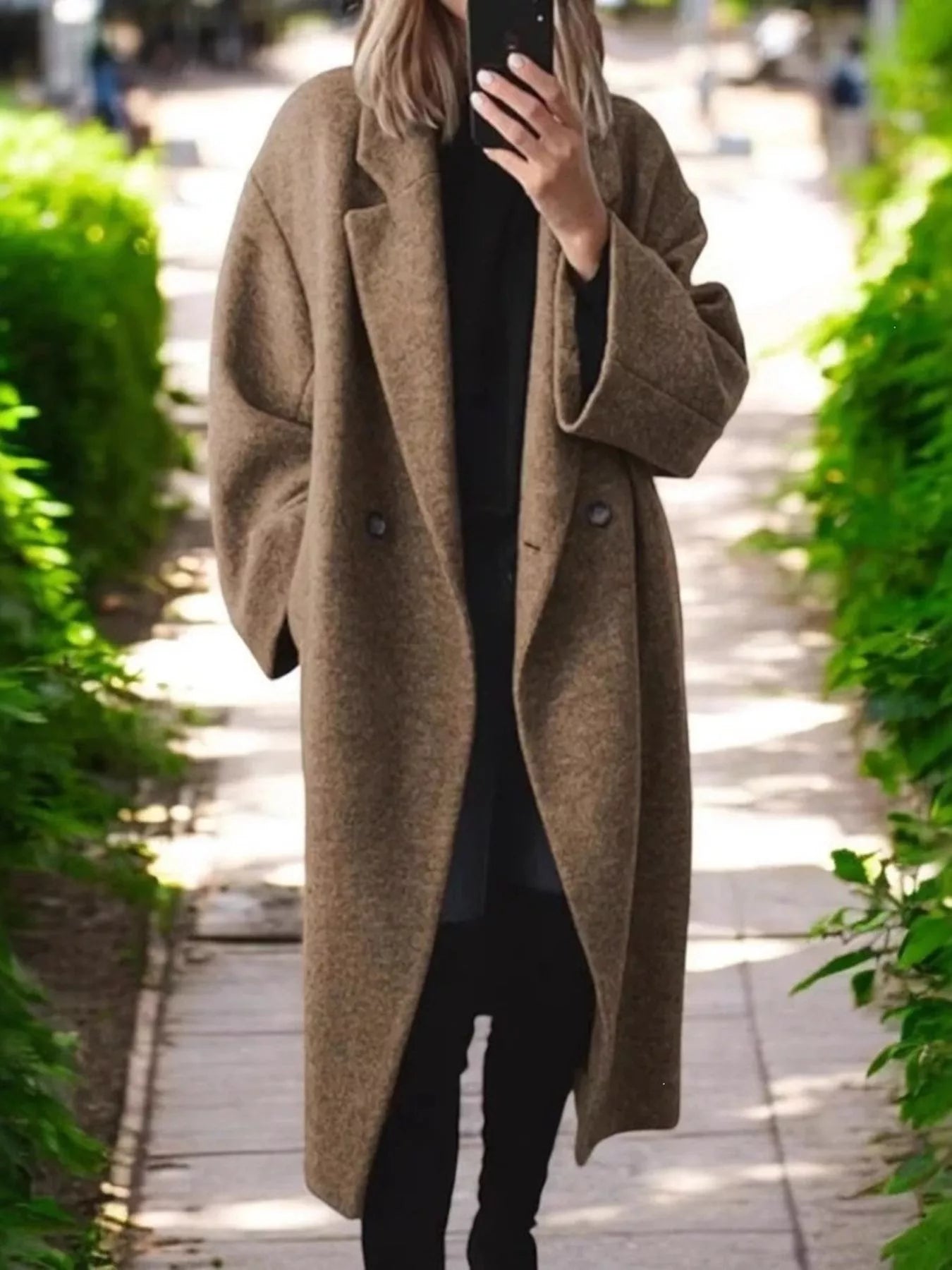 A woman takes a mirror selfie wearing a long, double-breasted brown wool-blend coat over a black outfit, standing on a paved pathway lined with green bushes.