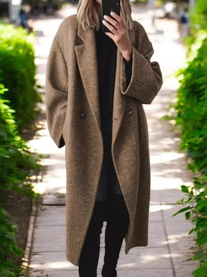 A woman takes a mirror selfie wearing a long, double-breasted brown wool-blend coat over a black outfit, standing on a paved pathway lined with green bushes.