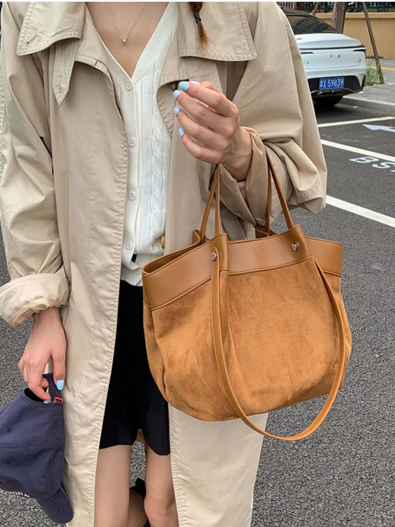 A person wearing a beige trench coat and white blouse carries a large brown suede-like shoulder bag with a long strap in an outdoor street setting.