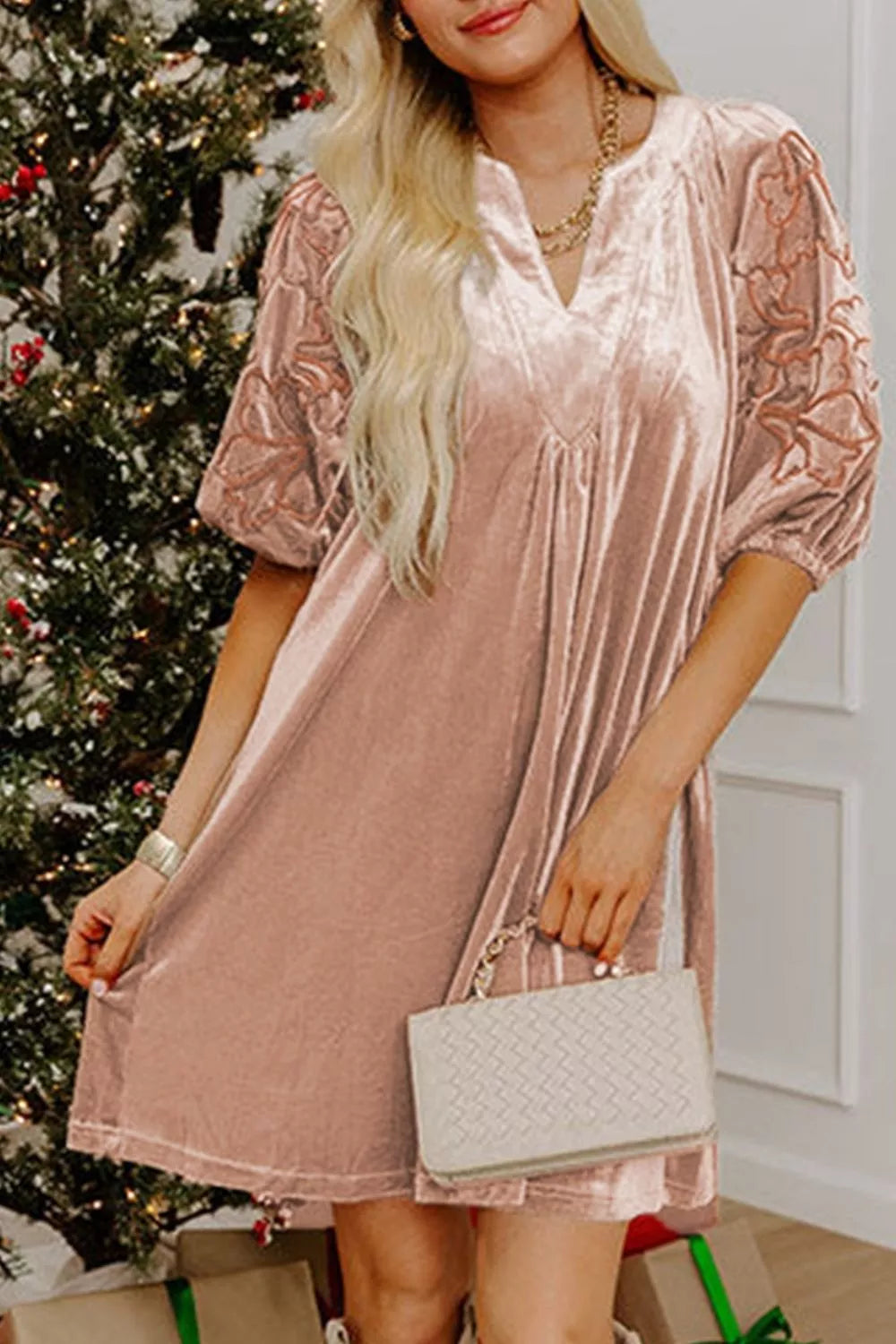 A woman wearing a dusty pink velvet mini dress with sheer lace puff sleeves, holding a white quilted handbag in front of a decorated Christmas tree.