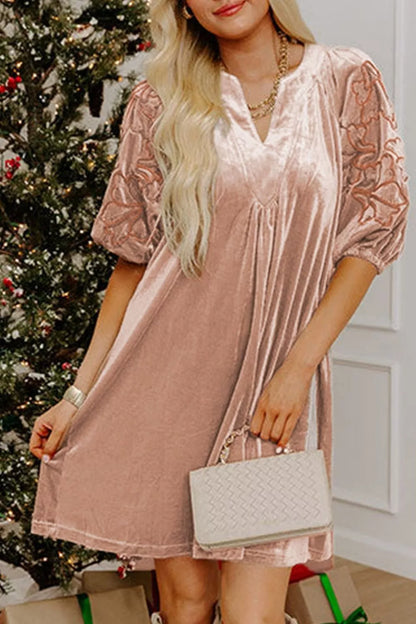 A woman wearing a dusty pink velvet mini dress with sheer lace puff sleeves, holding a white quilted handbag in front of a decorated Christmas tree.