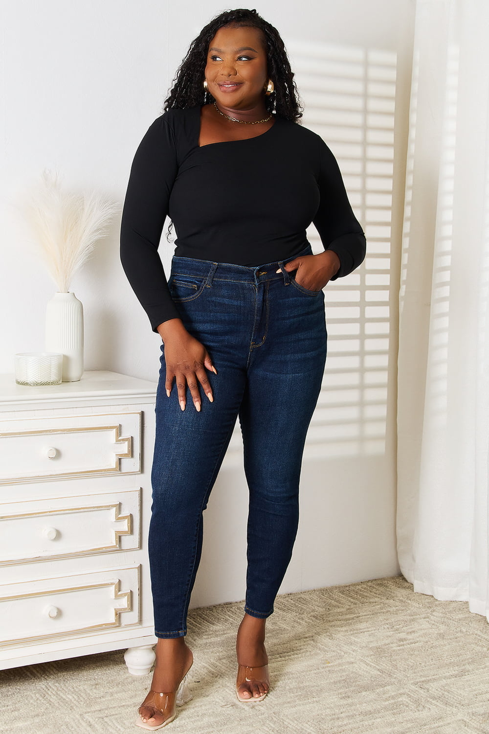 Woman wearing a black top and blue jeans standing in a room with white walls and a white dresser.