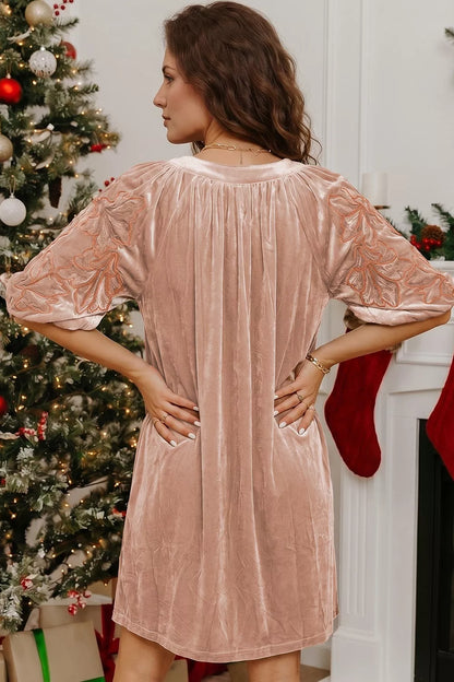 A woman wearing a dusty rose velvet holiday dress with short puff sleeves and side pockets, standing in front of a decorated Christmas tree and fireplace.