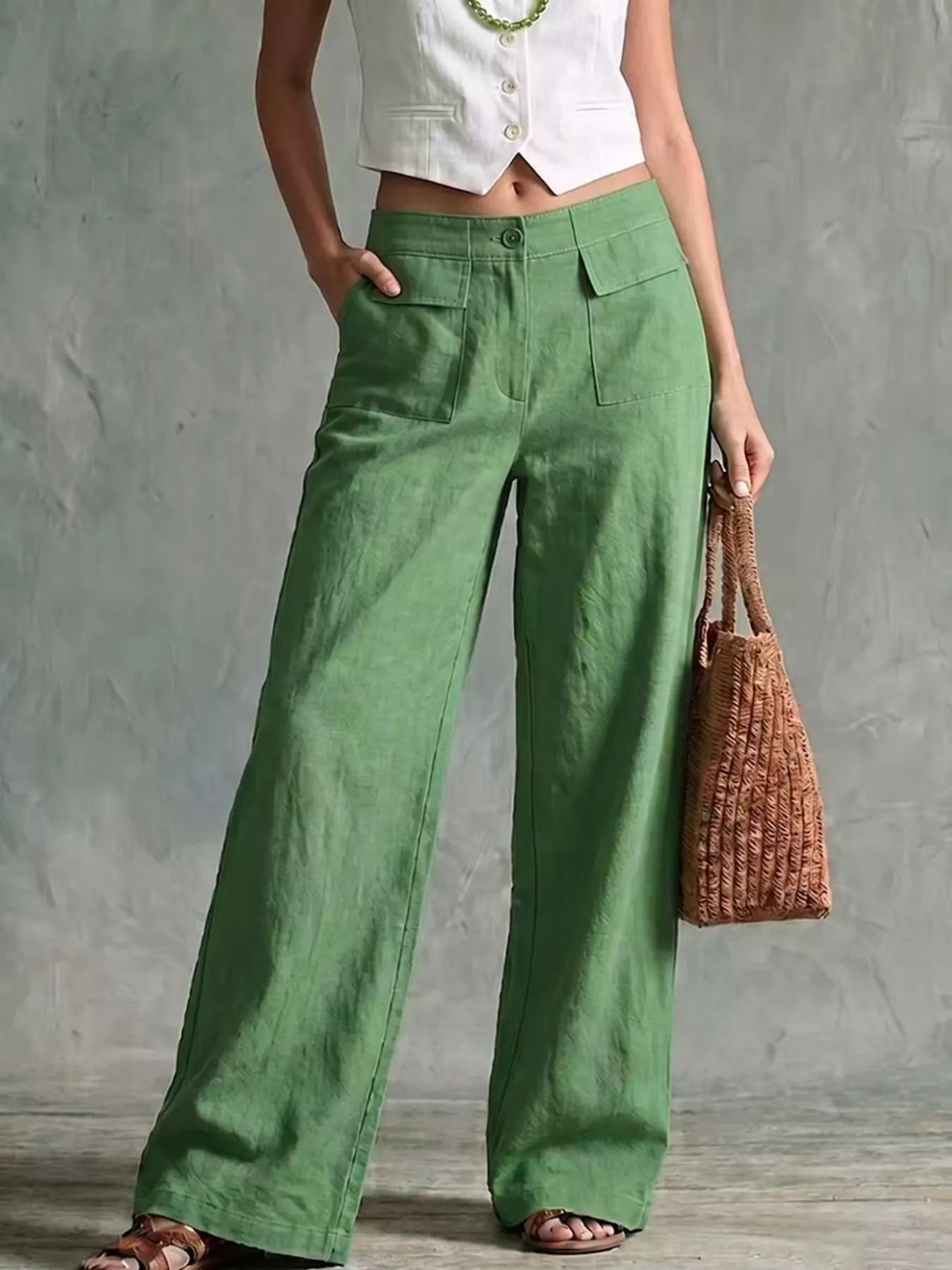 A model wearing green wide-leg linen pants, styled with a white cropped vest, a green beaded necklace, and brown sandals, holding a brown woven tote bag against a textured gray wall.