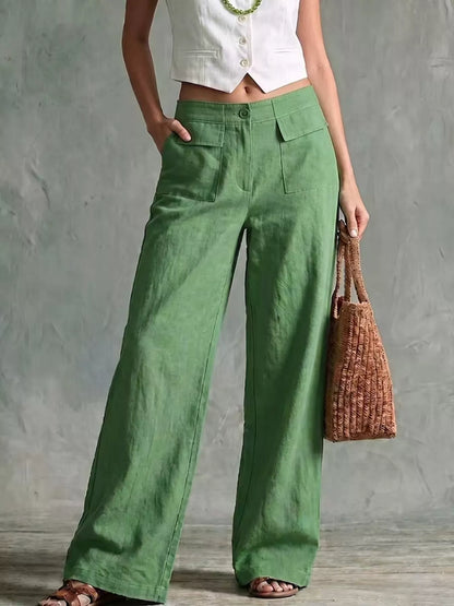 A model wearing green wide-leg linen pants, styled with a white cropped vest, a green beaded necklace, and brown sandals, holding a brown woven tote bag against a textured gray wall.