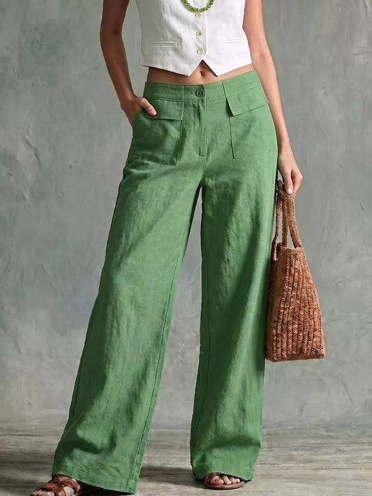 A model wearing green wide-leg linen pants, styled with a white cropped vest, a green beaded necklace, and brown sandals, holding a brown woven tote bag against a textured gray wall.