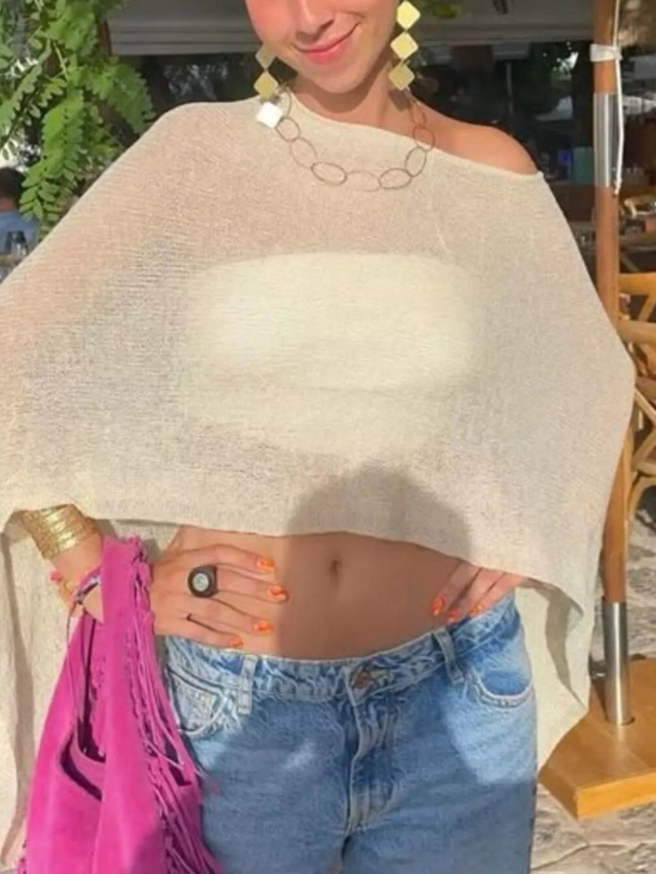 A woman wearing a light beige knit poncho-style top with blue jeans, gold statement earrings, and a gold chain necklace, holding a bright pink fringed bag in an outdoor setting.