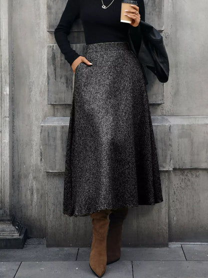 Person wearing a black top and gray skirt with brown boots against a gray wall.