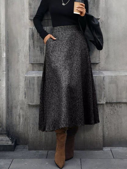 Person wearing a black top and gray skirt with brown boots against a gray wall.