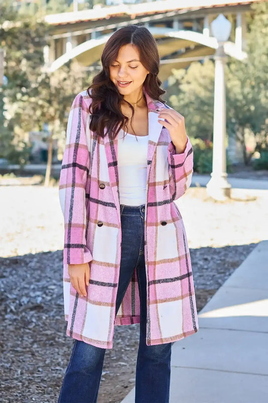 A woman wearing a pink and white plaid double-breasted coat over a white top and dark blue jeans, standing outdoors on a paved walkway near a covered patio.