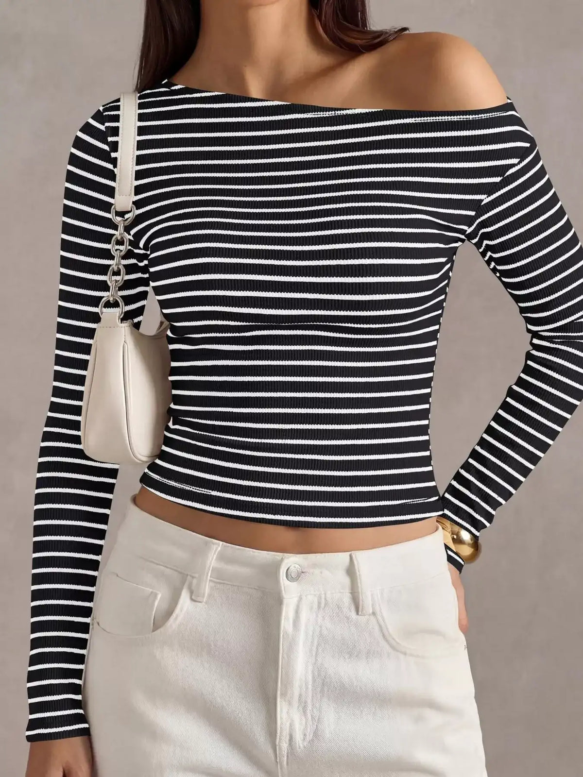 A woman wearing a black and white horizontally striped off-the-shoulder long-sleeve crop top, paired with white pants and carrying a small white shoulder bag.