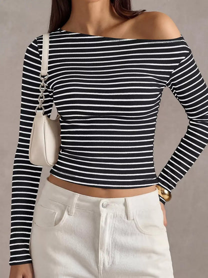 A woman wearing a black and white horizontally striped off-the-shoulder long-sleeve crop top, paired with white pants and carrying a small white shoulder bag.