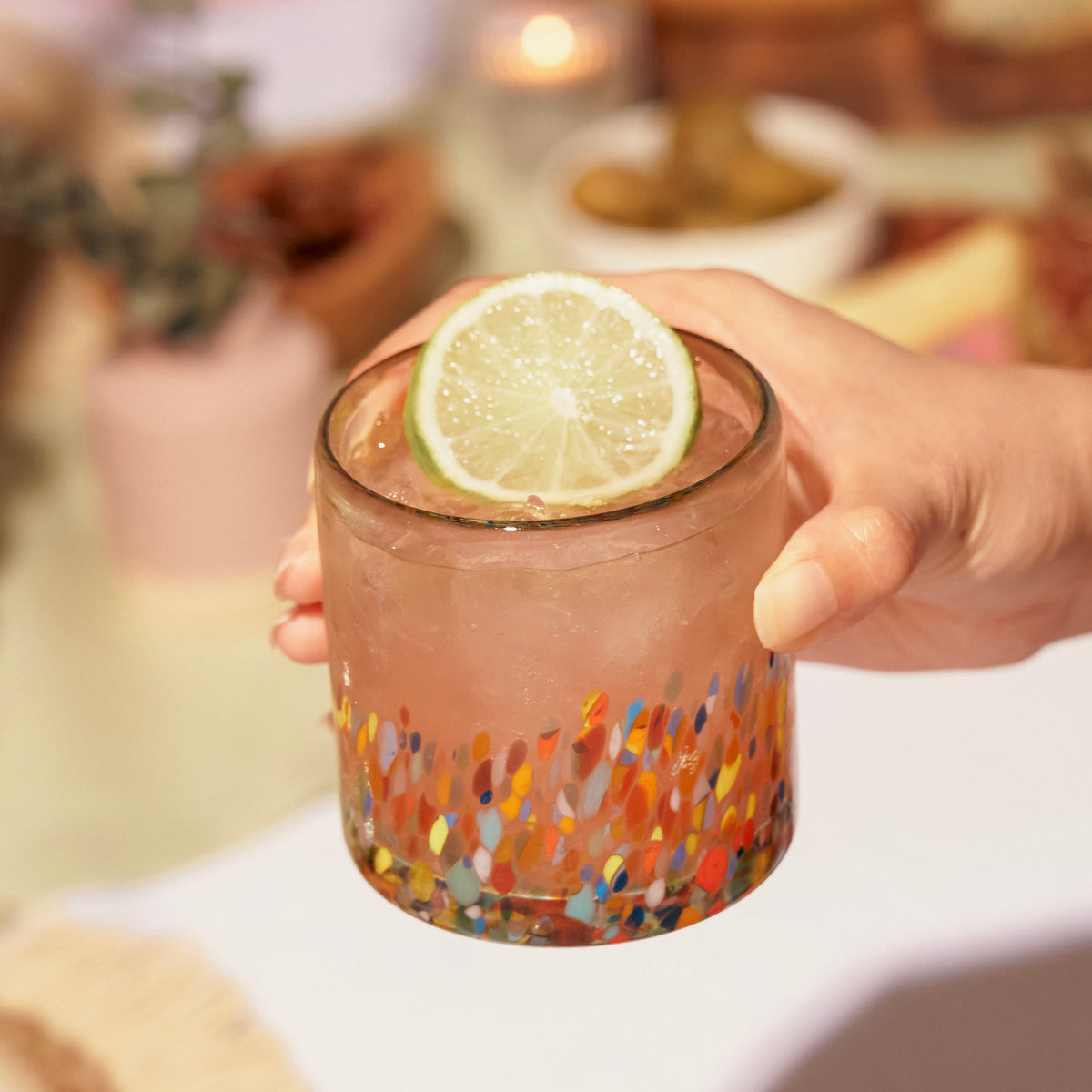 Hand holding a colorful speckled glass tumbler filled with an iced pink drink and a lime slice.