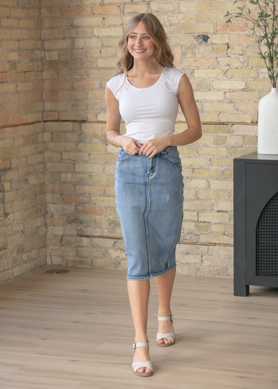 Woman wearing a white top and blue denim skirt standing in a room with a stone wall.