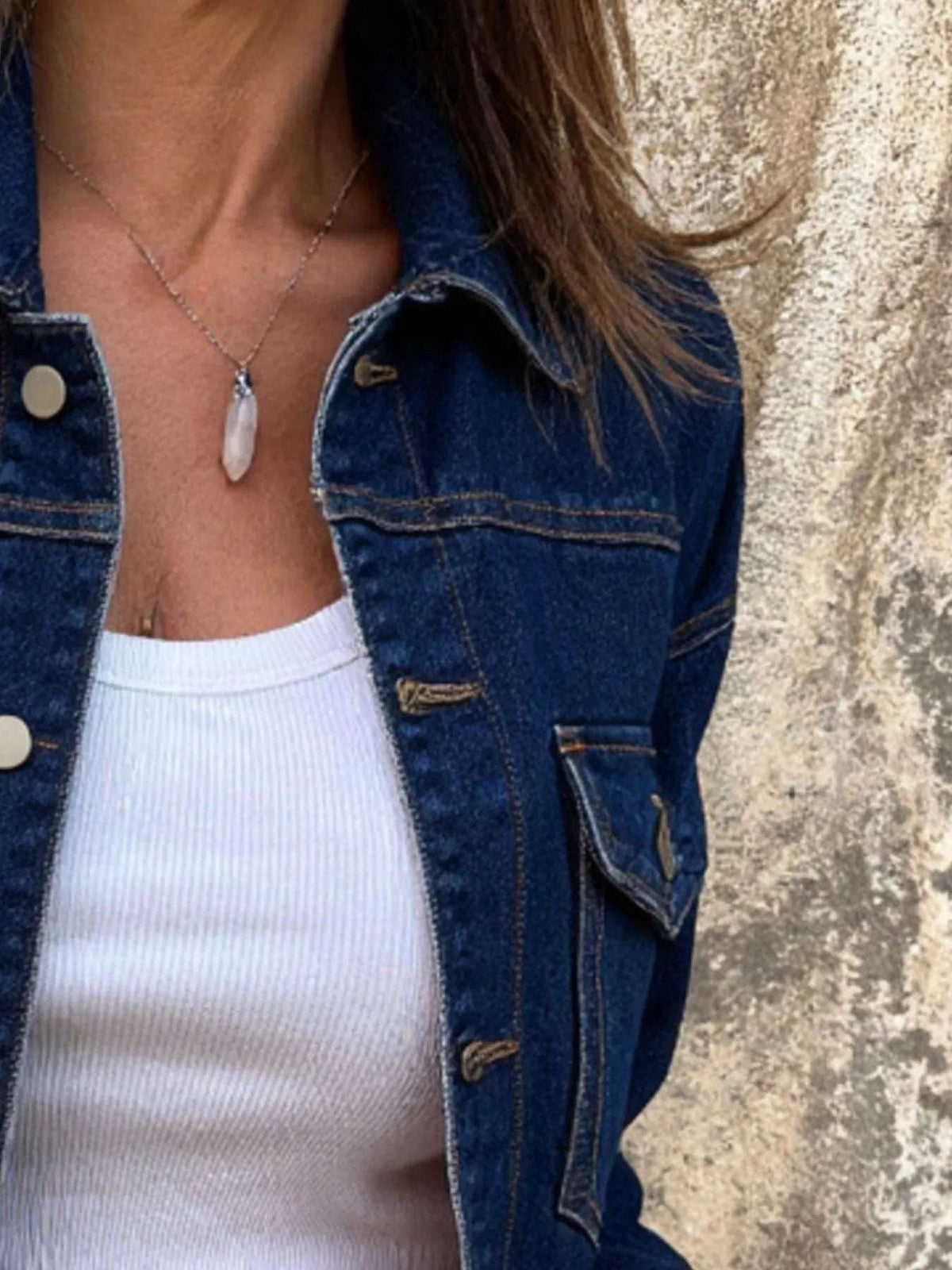 Person wearing a blue denim jacket over a white top against a textured wall.