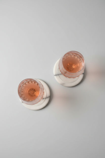 Two clear stemmed drinking glasses viewed from below, resting on a round woven placemat with blue, green, and beige stripes. The lower portions of the glasses show pinkish liquid at the top.
