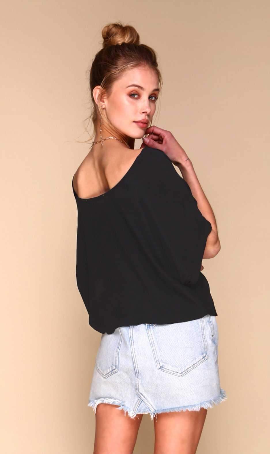 A woman wearing a black off-the-shoulder knit top and light-wash denim shorts, photographed against a plain beige studio background.