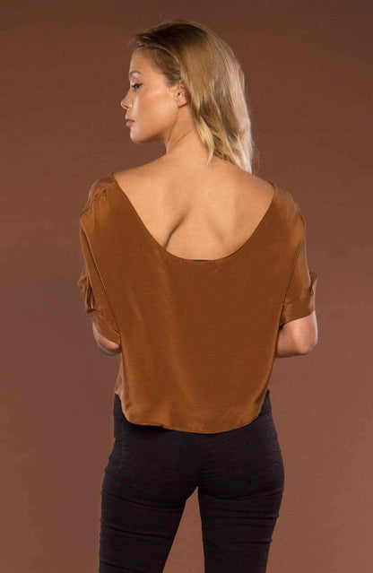 A woman shown from the back wearing a brown short-sleeve top with a wide, open low neckline and dark pants, standing against a solid brown studio background.