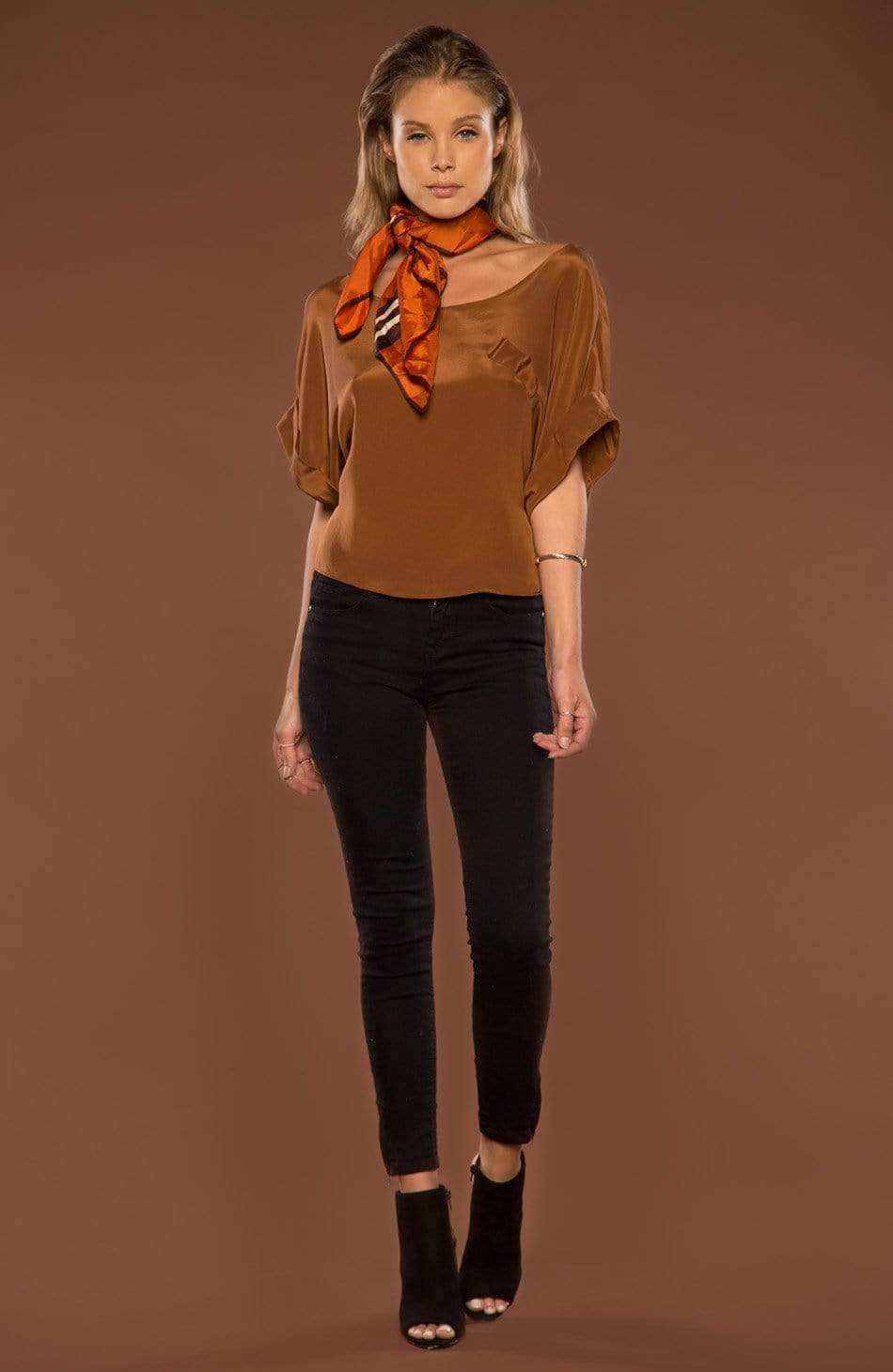 A woman modeling a brown short-sleeve top with a draped neckline, paired with black skinny pants and black ankle boots, accessorized with an orange patterned neck scarf, against a solid brown studio background.