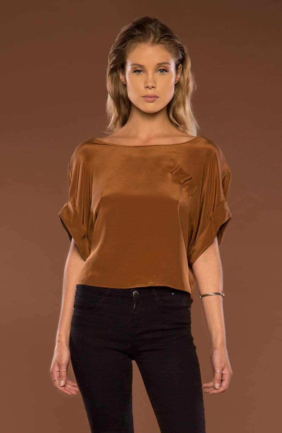 A woman modeling a brown wide-neck top with draped, short sleeves and black pants, set against a solid brown studio background.