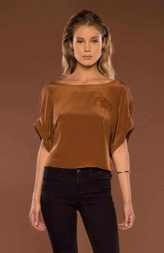 A woman modeling a brown wide-neck top with draped, short sleeves and black pants, set against a solid brown studio background.