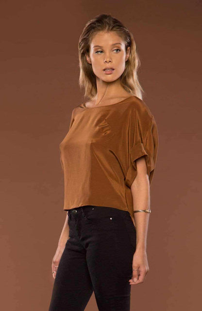 A woman modeling a brown short-sleeve top with a wide boat neckline and black pants, standing against a solid brown studio background.