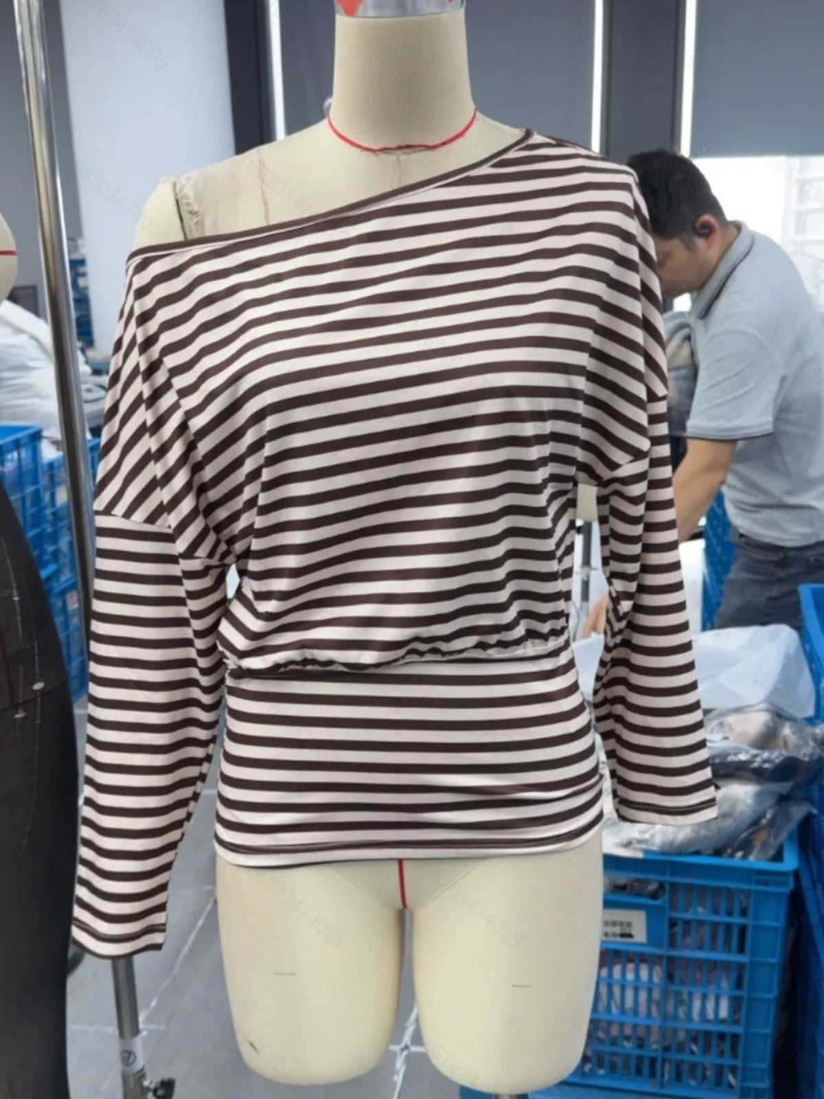 A long-sleeve off-the-shoulder top with horizontal brown and beige stripes, displayed on a beige mannequin torso in a warehouse or tailoring shop setting.