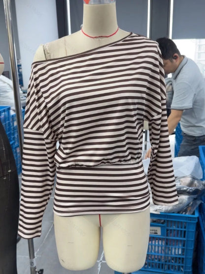 A long-sleeve off-the-shoulder top with horizontal brown and beige stripes, displayed on a beige mannequin torso in a warehouse or tailoring shop setting.