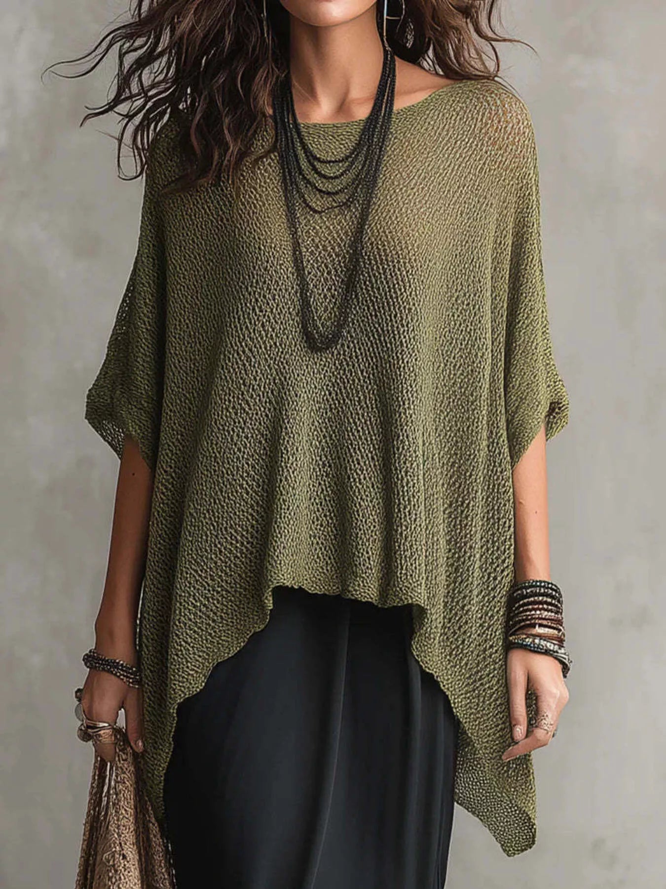 A woman wearing a loose, olive green open-knit poncho-style top with short wide sleeves and an asymmetrical hem, paired with black pants and layered dark necklaces, against a neutral gray background.