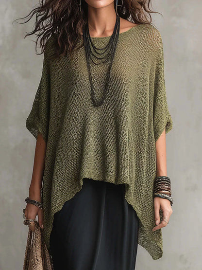 A woman wearing a loose, olive green open-knit poncho-style top with short wide sleeves and an asymmetrical hem, paired with black pants and layered dark necklaces, against a neutral gray background.
