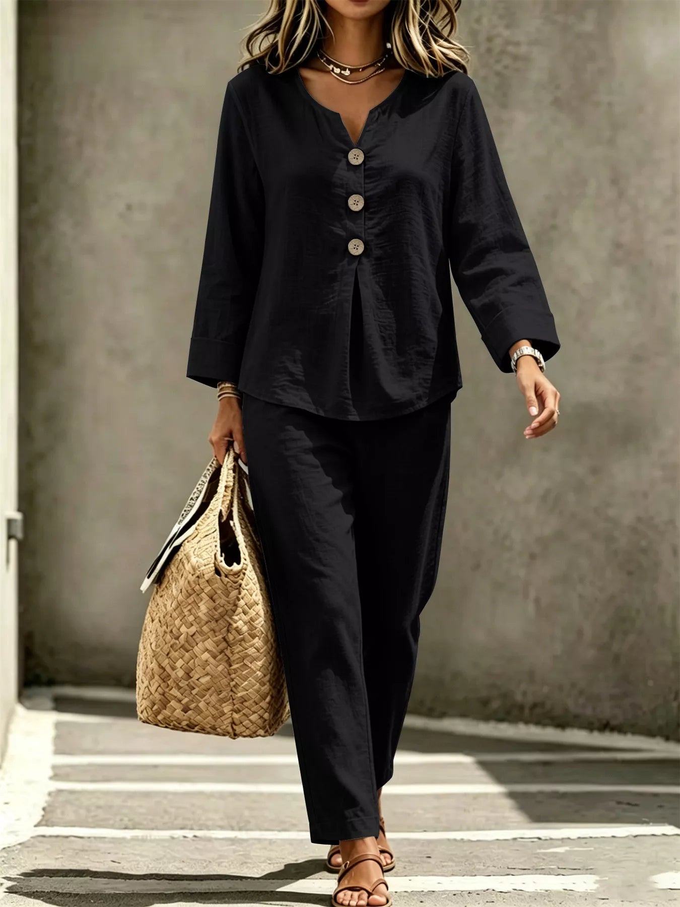 A woman wearing a black two-piece linen set consisting of a long-sleeve button-up top and matching trousers, carrying a woven straw tote bag.