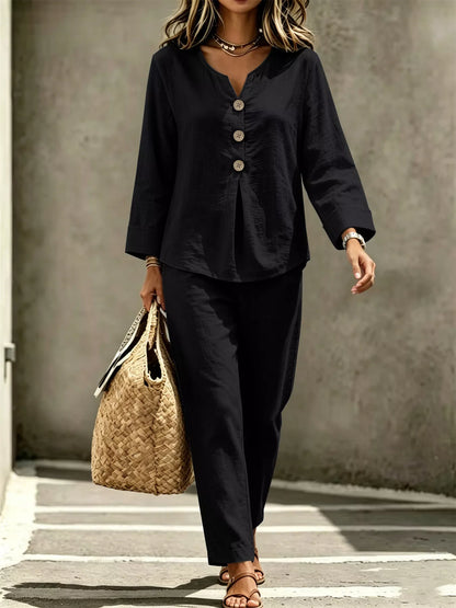 A woman wearing a black two-piece linen set consisting of a long-sleeve button-up top and matching trousers, carrying a woven straw tote bag.