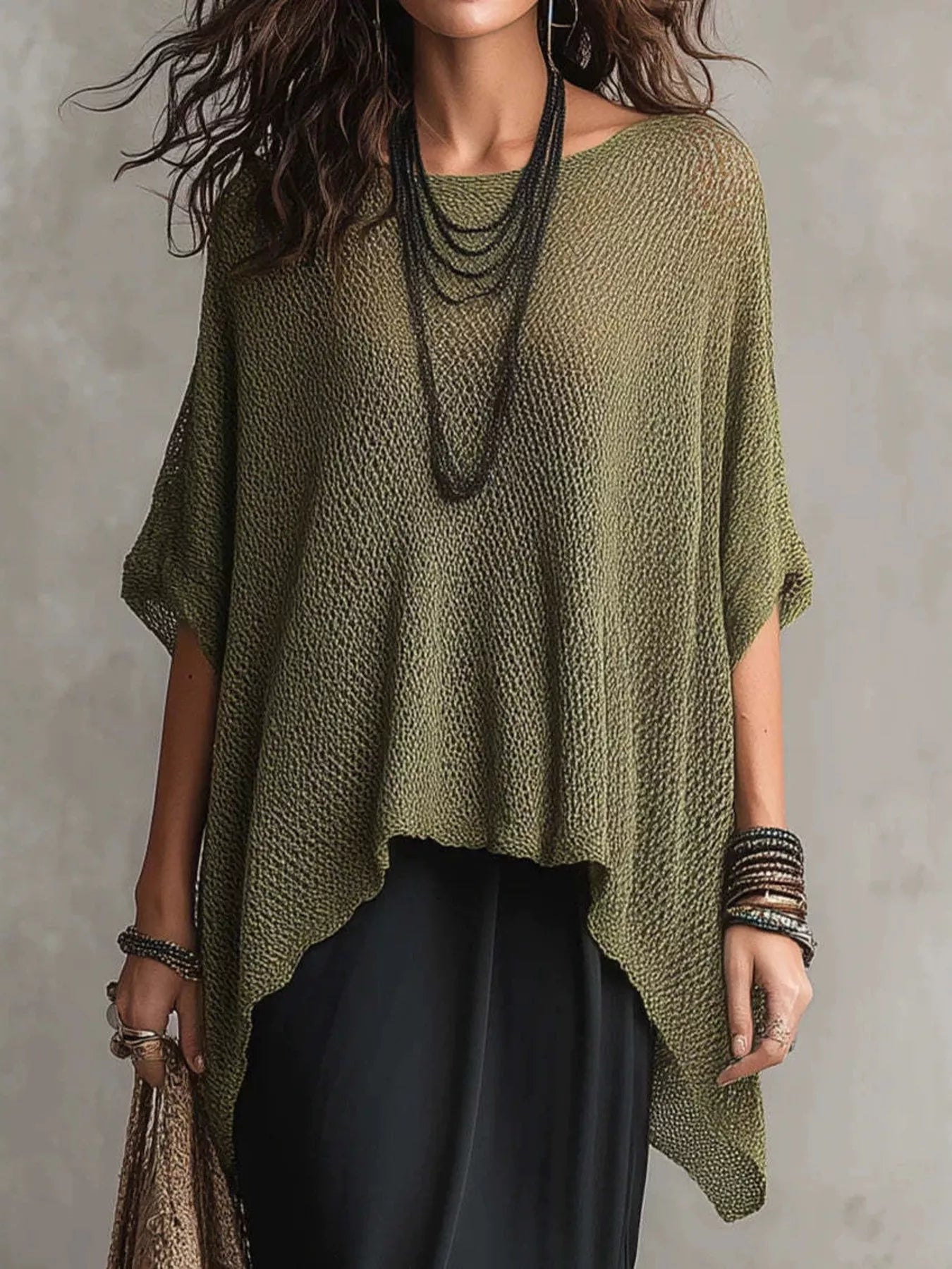 A woman wearing a loose, olive green open-knit poncho-style top with short wide sleeves, layered with long dark necklaces and bracelets, paired with black bottoms against a textured gray wall.