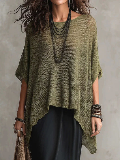 A woman wearing a loose, olive green open-knit poncho-style top with short wide sleeves, layered with long dark necklaces and bracelets, paired with black bottoms against a textured gray wall.