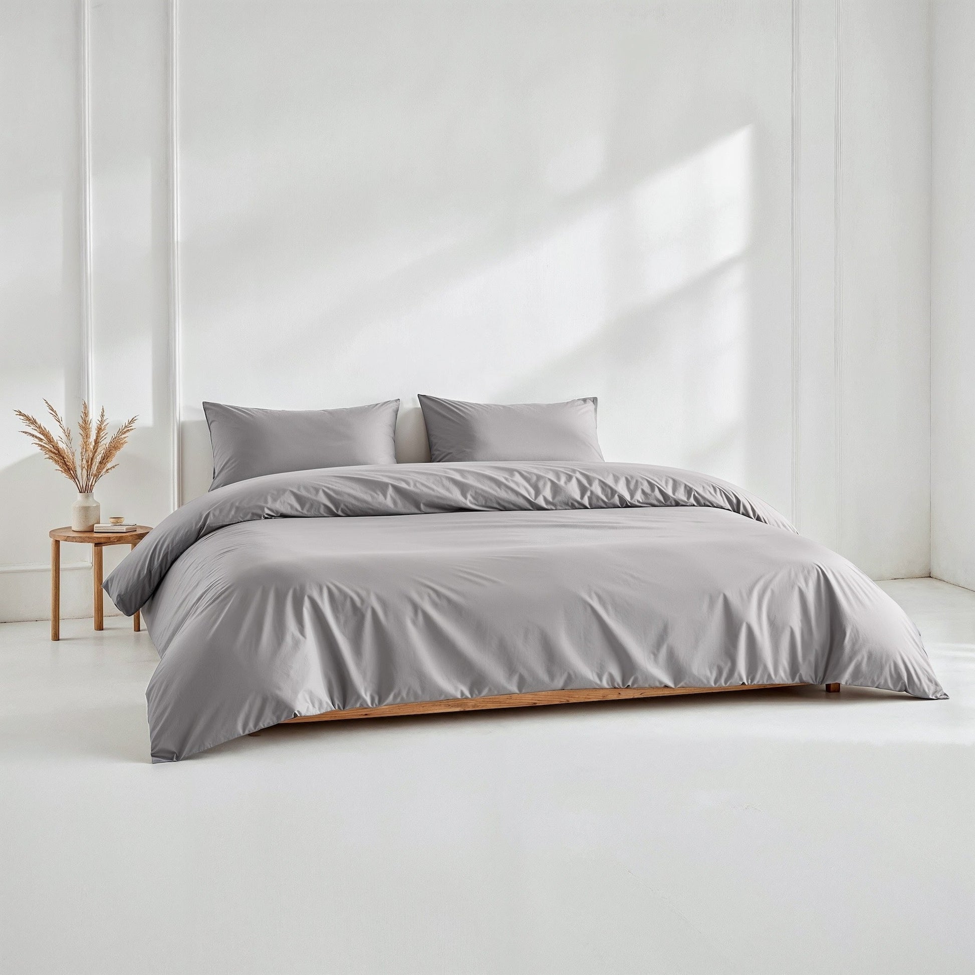 Gray bedding set on a bed in a minimalistic room with a white wall and wooden side table.