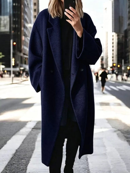 A woman takes a mirror selfie on a city crosswalk wearing a long, oversized navy blue wool coat over a black outfit.