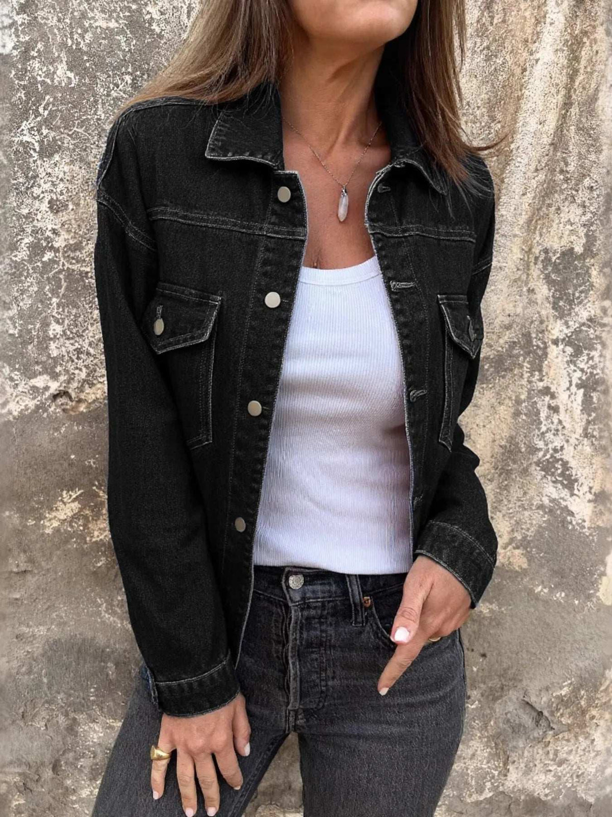 Person wearing a black denim jacket over a white top and dark jeans against a textured wall.