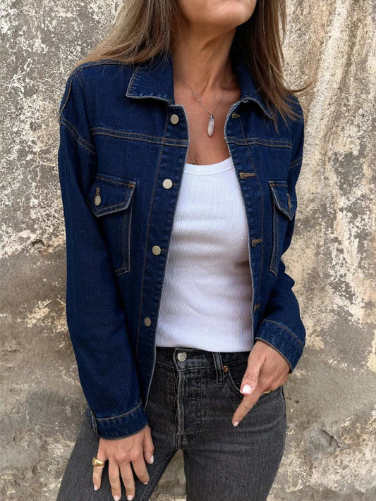 Person wearing a blue denim jacket over a white top and black jeans against a textured wall.