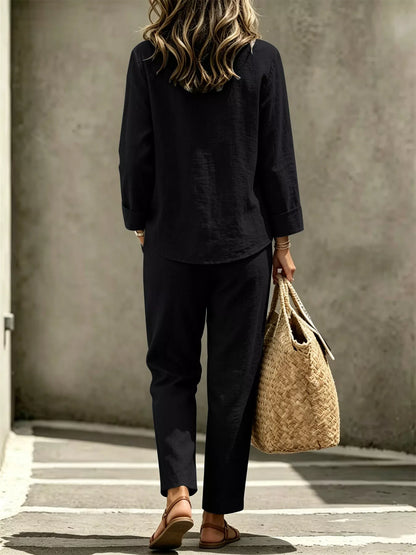 A woman viewed from behind wearing a black linen-blend long-sleeve top and matching black trousers, carrying a large woven straw tote bag against a textured concrete wall.