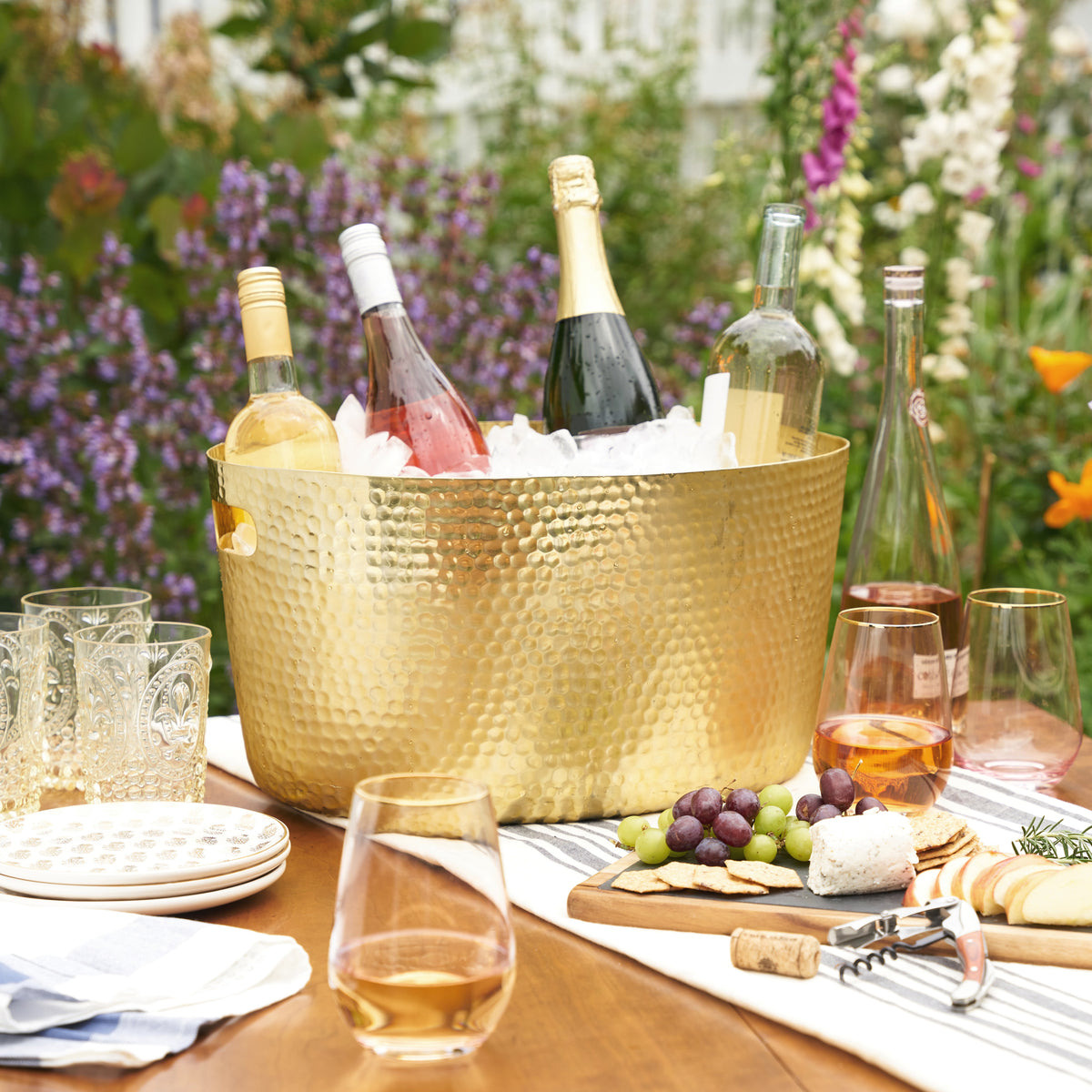 Hammered gold beverage tub filled with ice and wine bottles on an outdoor table beside glassware and a cheese board.