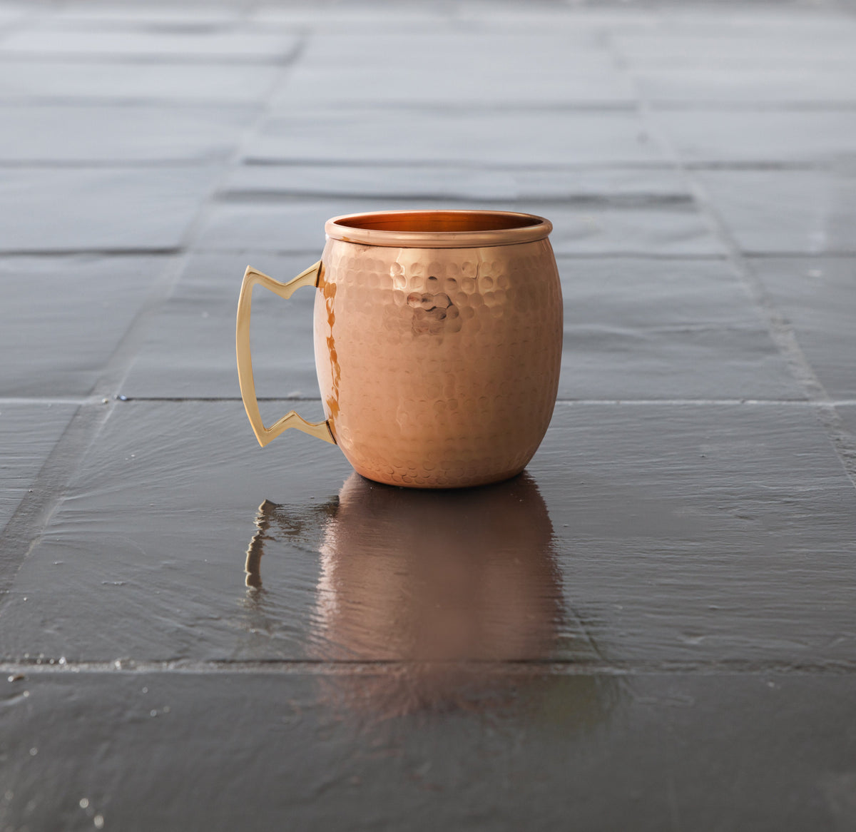 Hammered copper mule mug on a dark reflective tiled surface, with the handle facing left.