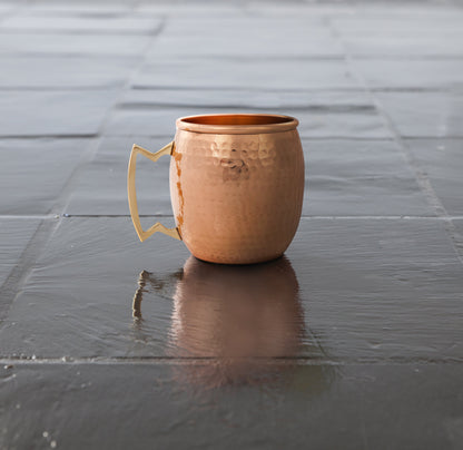 Hammered copper mule mug on a dark reflective tiled surface, with the handle facing left.