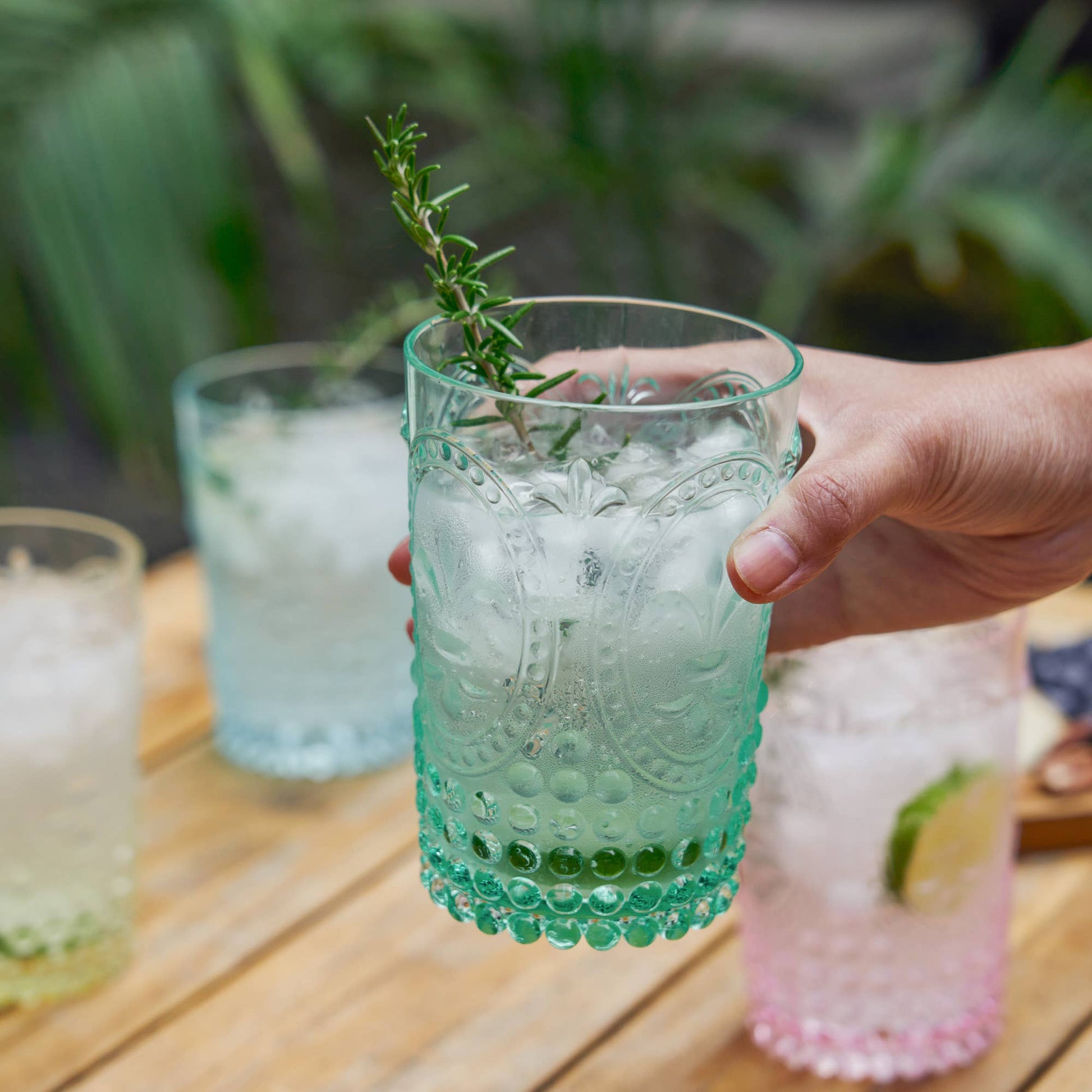Hand holding a decorative glass with a drink and ice cubes on a wooden table.