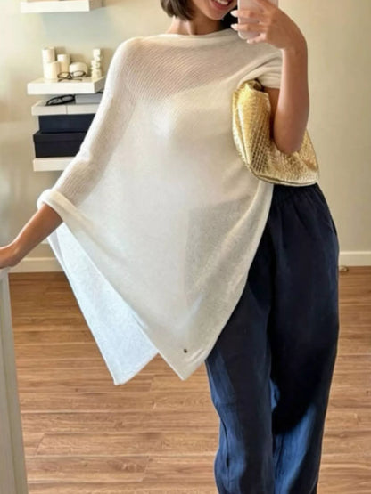 A woman wearing a cream-colored knit poncho-style top and dark blue wide-leg pants, holding a gold textured clutch bag in an indoor setting with wooden floors.