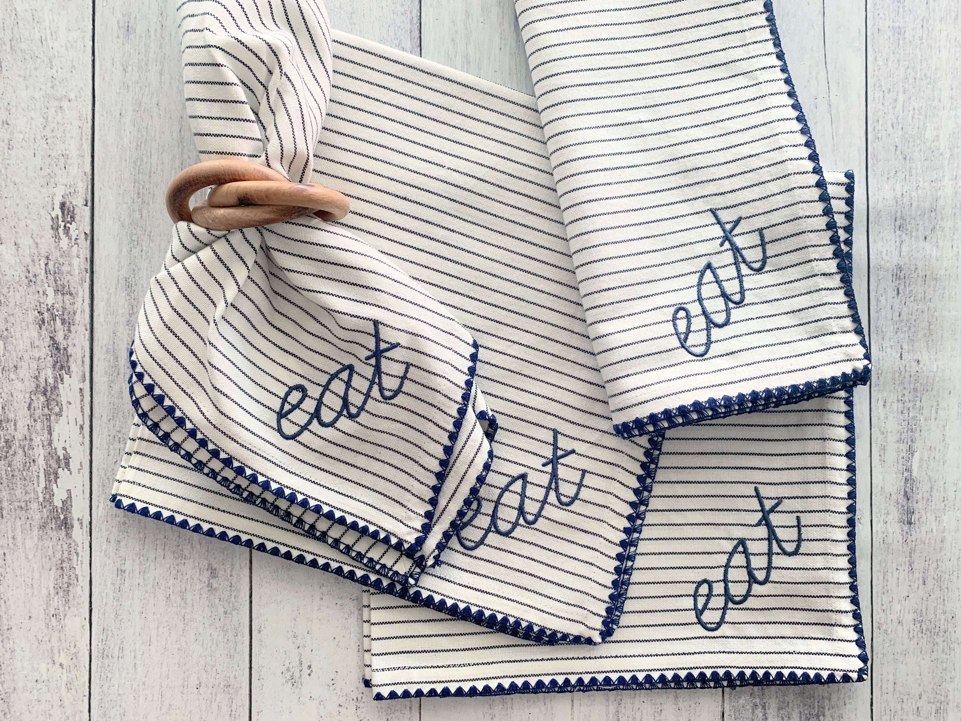 Four striped fabric napkins with blue eat embroidery and one round wood napkin ring on a white wood surface.