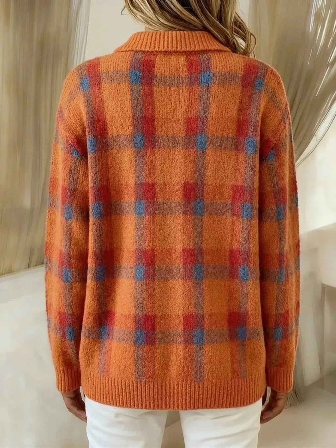 A person shown from the back wearing a fuzzy orange knit sweater with a red and blue plaid pattern, paired with white pants in an indoor setting.