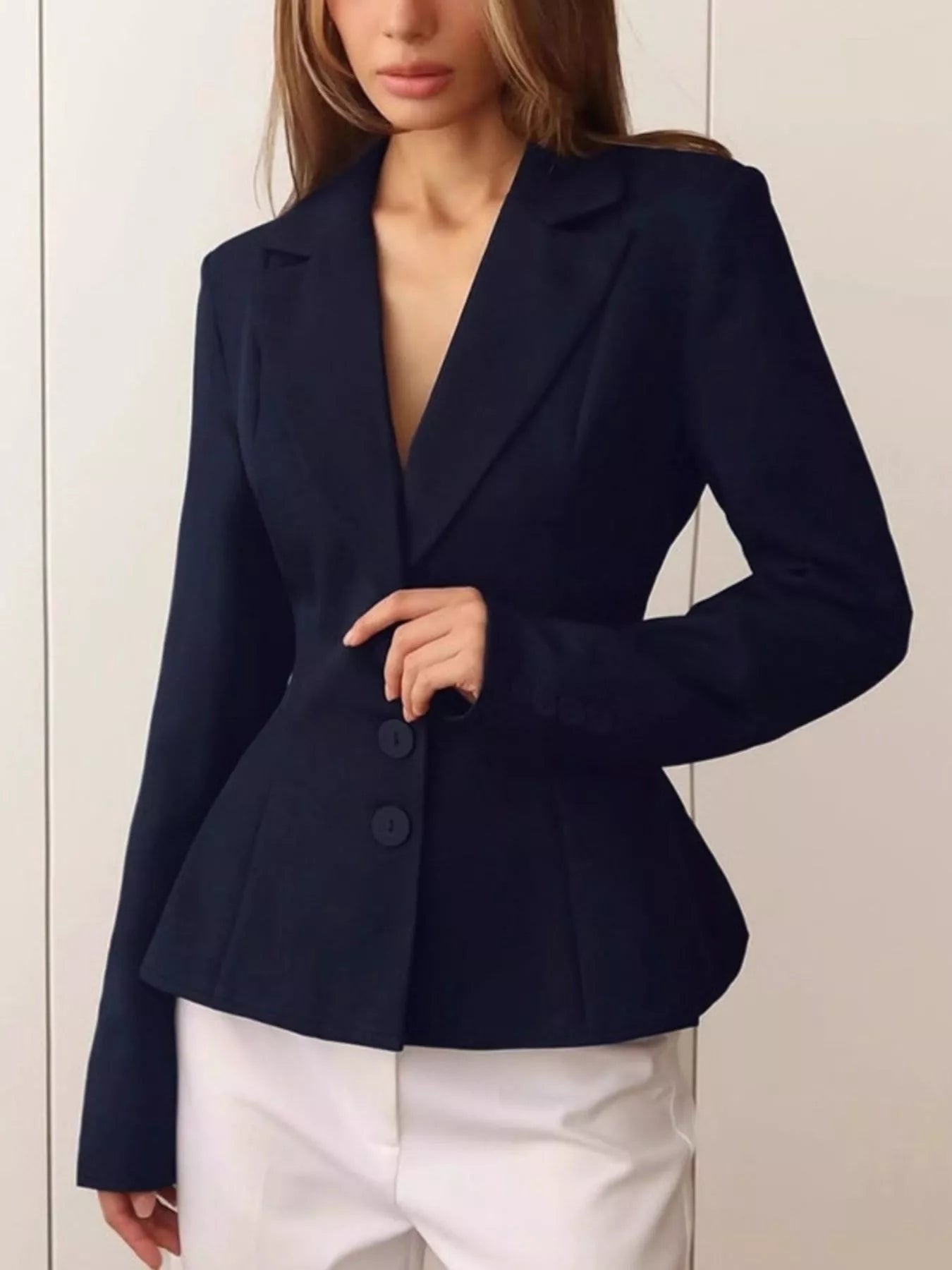 A woman wearing a dark navy blue tailored blazer with a deep V-neck and three front buttons, paired with white pants, shown against a light neutral paneled wall.