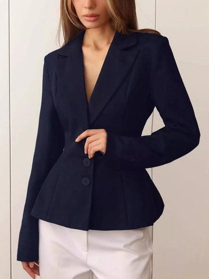 A woman wearing a dark navy blue tailored blazer with a deep V-neck and three front buttons, paired with white pants, shown against a light neutral paneled wall.