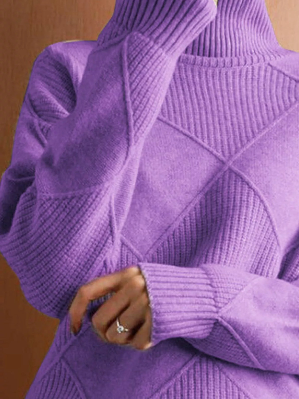 A close-up of a person wearing a chunky purple knit turtleneck sweater with diamond-shaped textured panels and ribbed cuffs, against a warm brown background.